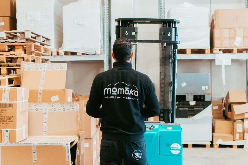 Un operatore logistico Momoka, di schiena, mentre utilizza un transpallet elettrico per spostare in modo rapido e sicuro un bancale pieno di scatole di cartone all'interno del magazzino.