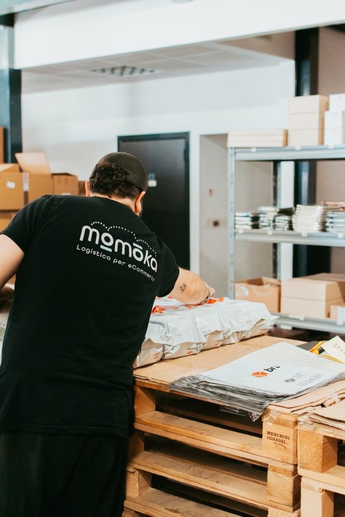 Operatore Momoka al lavoro in magazzino con t-shirt aziendale, impegnato nella gestione degli ordini.