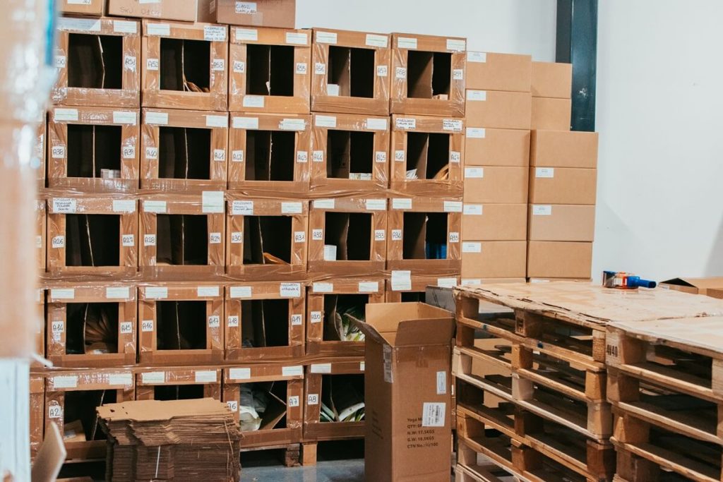 Dettaglio di scatole di cartone modificate, impilate e codificate con etichette alfanumeriche per fungere da postazioni di picking manuale, facilitando l'inventario e la preparazione degli ordini.