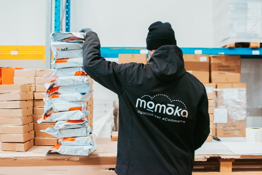 Un operatore logistico Momoka visto di schiena, con una giacca nera con logo, mentre impila ordinatamente una pila alta di pacchi confezionati e pronti per la spedizione, assicurando che siano pronti per la consegna.