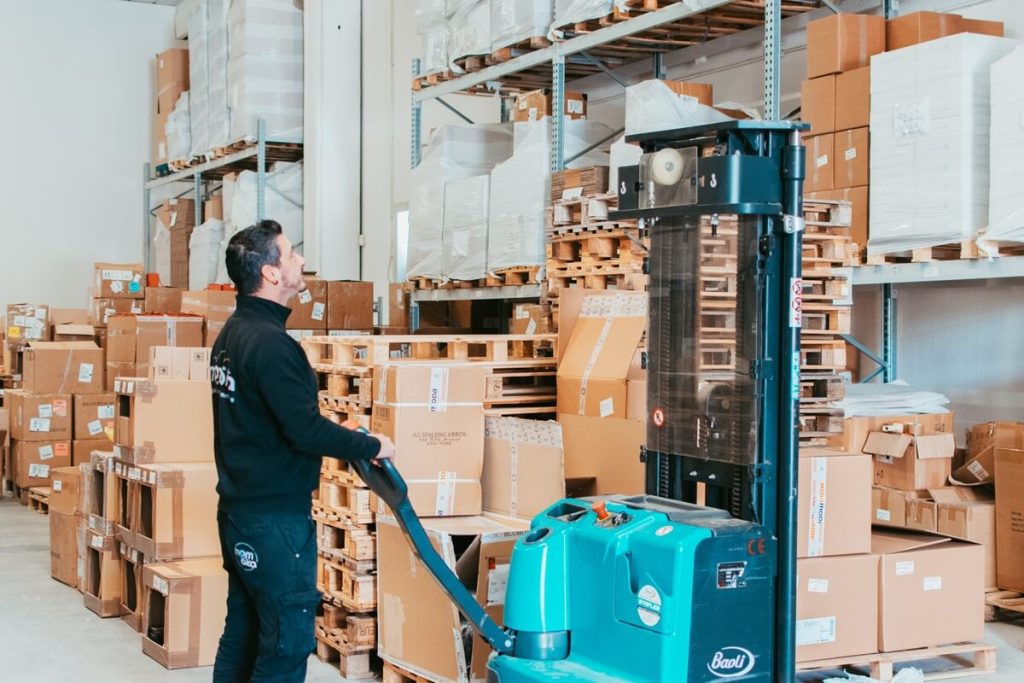 Un operatore logistico Momoka mentre utilizza un transpallet elettrico per spostare in modo rapido e sicuro un bancale pieno di scatole di cartone all'interno del magazzino, garantendo l'efficienza dei flussi di magazzino.