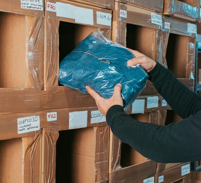 Un primo piano dettagliato di una mano che posiziona un articolo (un maglione blu) confezionato in un contenitore a scaffale etichettato, mostrando un momento chiave dell'operazione di prelievo o imballaggio Mago Magazzino Momoka.